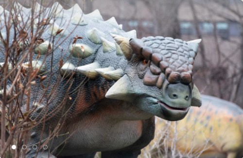 A realistic dinosaur sculpture with a spiky back, surrounded by bare branches and a blurred background.