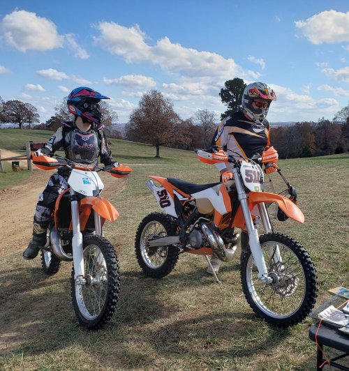 Two riders in motocross gear stand beside their orange dirt bikes on a grassy field under a blue sky with clouds.