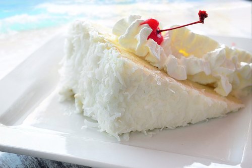 A slice of coconut cream cake topped with whipped cream and a cherry, served on a white plate.