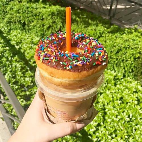 A hand holding a large donut topped with chocolate icing and colorful sprinkles, placed on a coffee cup.