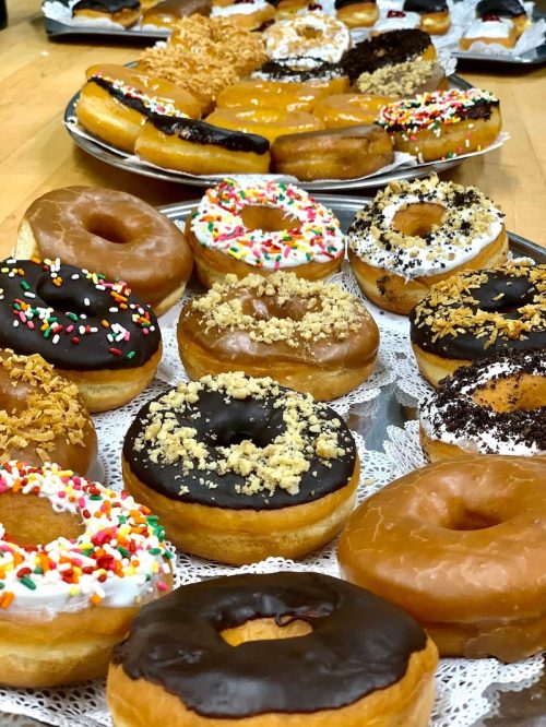 A variety of colorful donuts on trays, featuring sprinkles, chocolate, and cream toppings.
