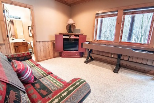 Cozy room with a patterned couch, a TV cabinet, and a keyboard, featuring large windows overlooking a wooded area.