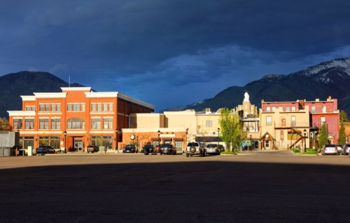 A scenic view of a small town with buildings and mountains under a dramatic sky. Cars are parked in the foreground.