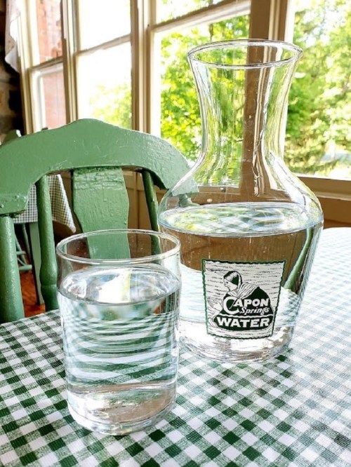 A glass of water next to a clear pitcher on a checkered tablecloth, with sunlight streaming through a window.