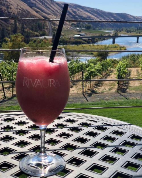 A refreshing pink drink in a glass with a straw, set on a table overlooking a scenic vineyard and river view.