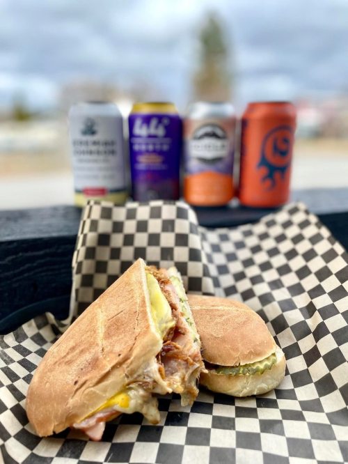 A sandwich with layers of meat and pickles on a checkered paper, with colorful cans of beverages in the background.