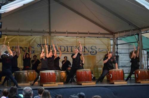 A group of performers in black attire play large drums on stage, showcasing a dynamic traditional music performance.