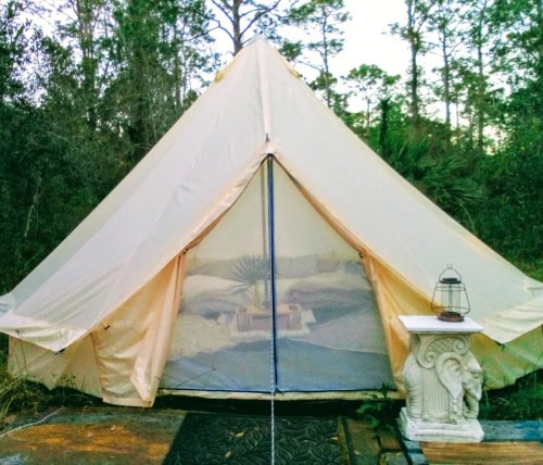 A cozy glamping tent with a mesh entrance, surrounded by trees and a decorative table beside it.