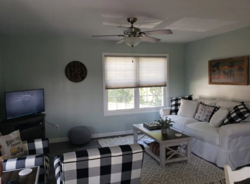 Cozy living room with a white sofa, checkered chairs, a coffee table, and a TV, featuring light blue walls and natural light.