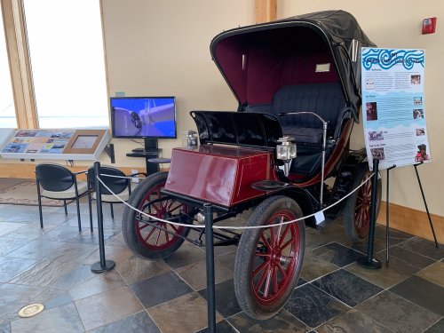A vintage car displayed indoors, with informational panels and a screen in the background.
