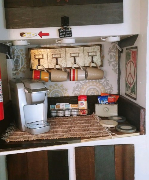Cozy coffee station with a Keurig, colorful mugs, and storage containers on a rustic shelf. Peace sign decor on the wall.