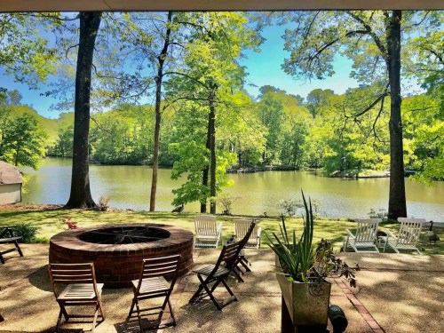 A serene lakeside view with a fire pit, surrounded by lush greenery and chairs on a sunny day.