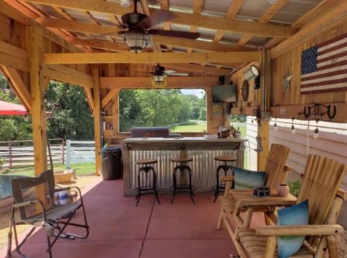 Cozy outdoor patio with wooden beams, seating, a bar area, and an American flag, surrounded by greenery.