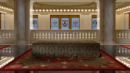 Elegant interior featuring a decorative bench on a patterned rug, with ornate windows and marble columns in the background.