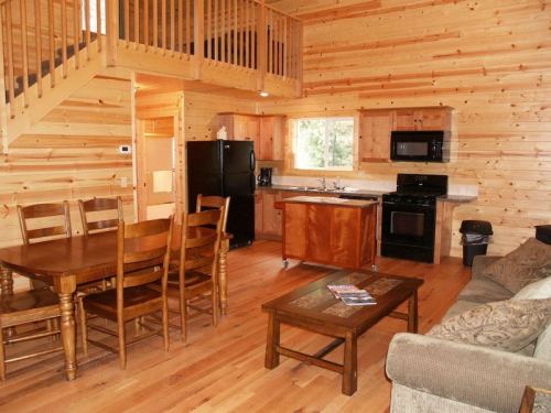 Cozy wooden cabin interior featuring a dining table, kitchen, and living area with a sofa and staircase.