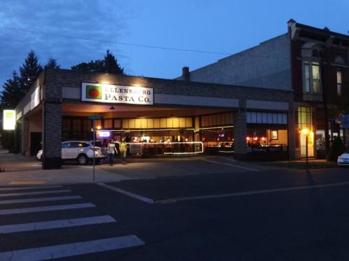 A pasta restaurant at dusk, with outdoor seating and cars parked nearby, set in a city street.
