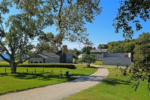 A scenic view of a gravel path leading through green grass, with historic buildings and trees under a clear blue sky.