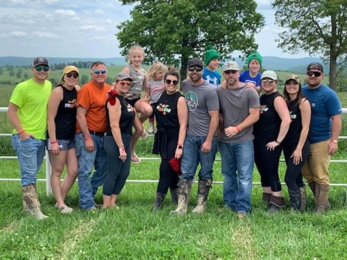 A group of people in casual attire and boots pose together outdoors, with green fields and hills in the background.