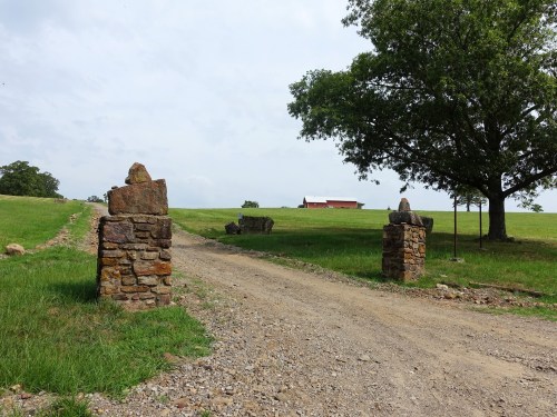 A gravel road leads through stone pillars to a distant red barn, surrounded by green fields and a large tree.