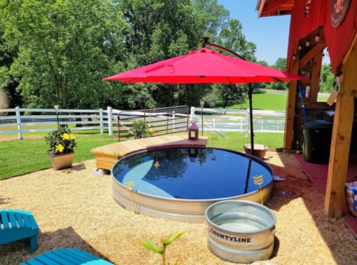 A circular pool with a red umbrella, surrounded by wooden decking and greenery, in a sunny outdoor setting.