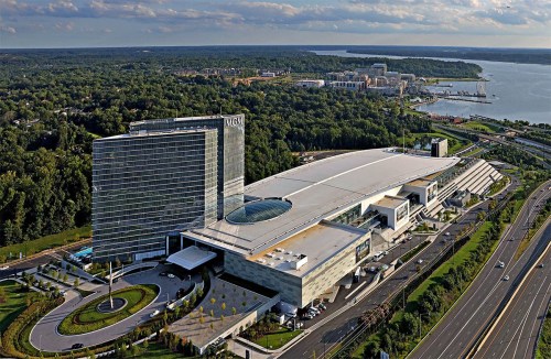 Aerial view of a modern hotel and casino complex near a lake, surrounded by greenery and a highway.