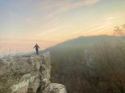 A person stands on the edge of a rocky cliff, overlooking a misty valley at sunrise.