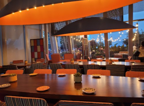 A modern restaurant interior with colorful seating, a large wooden table, and warm lighting, overlooking an outdoor area.