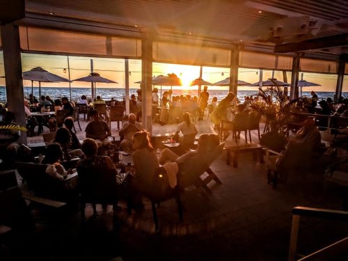 A lively beachside restaurant at sunset, filled with people enjoying drinks and the view.