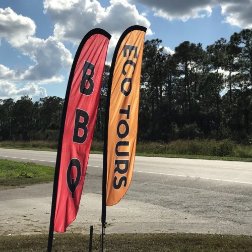 Two tall flags by the roadside: one red with "BBQ" and one orange with "ECO TOURS." Green trees in the background.