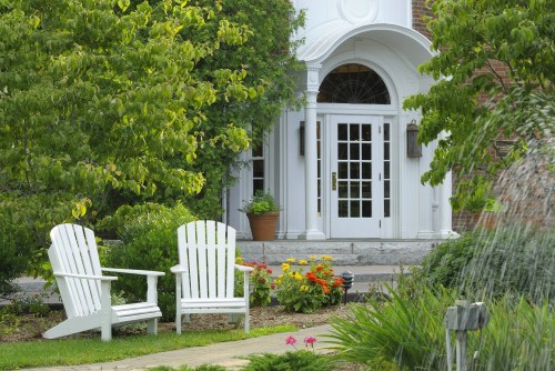 Two white Adirondack chairs sit in a garden with colorful flowers, facing a grand entrance with large double doors.