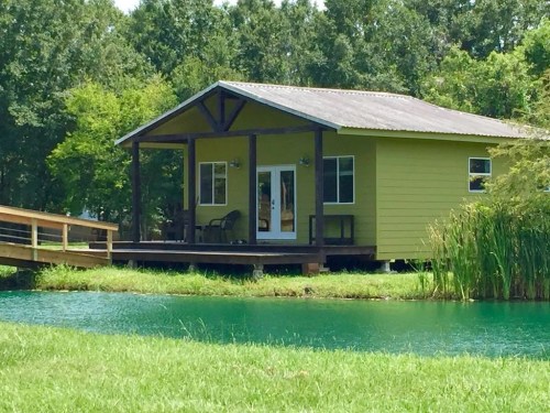 A small green cabin by a tranquil pond, surrounded by trees and greenery, with a wooden deck and chairs.