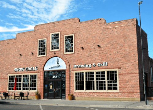 Exterior of Snow Eagle Brewing & Grill, a brick building with large windows and a sign, under a blue sky.