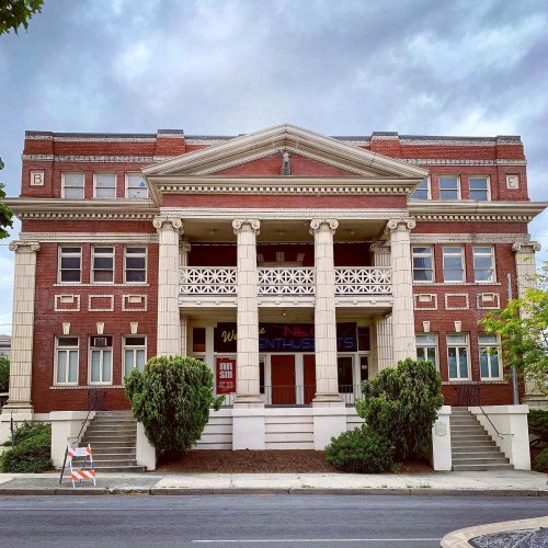 Historic brick building with columns, large windows, and greenery, under a cloudy sky.