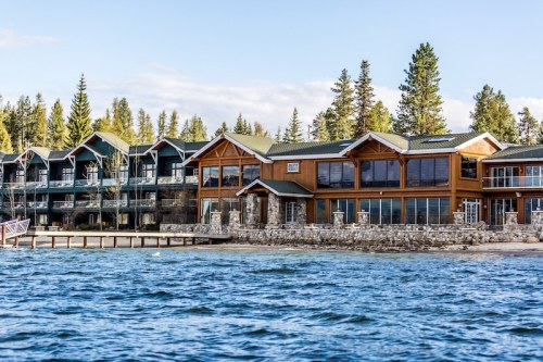 Lakeside view of a rustic lodge with stone accents and large windows, surrounded by trees and water.