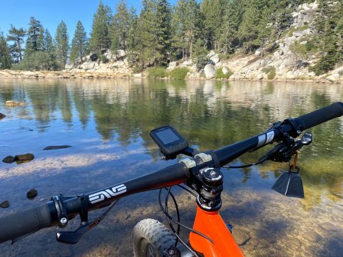 A close-up of a mountain bike handlebar near a calm lake surrounded by trees and rocky terrain.
