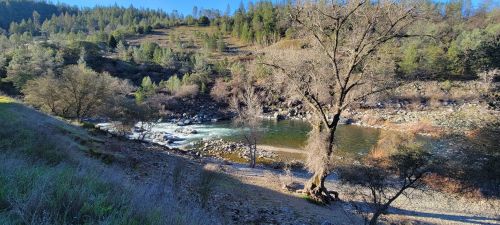 A serene river scene surrounded by trees and hills, with rocky banks and clear blue skies.