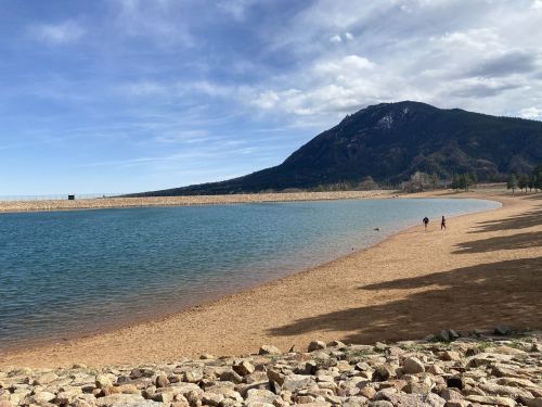 A serene lakeside scene with a sandy shore, mountains in the background, and two people walking along the water's edge.
