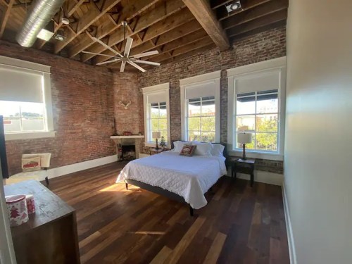 Bright bedroom with a wooden floor, exposed brick walls, large windows, and a cozy bed.