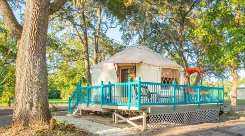 A cozy yurt with a wooden deck, surrounded by trees and greenery, featuring an orange chair on the porch.