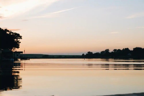 A serene sunset over a calm lake, reflecting soft colors and silhouettes of trees and buildings along the shore.