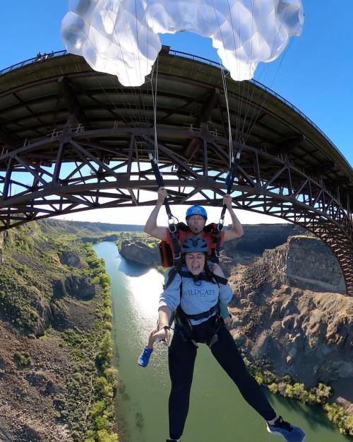 Two people tandem paragliding above a river, with a bridge in the background and a clear blue sky.