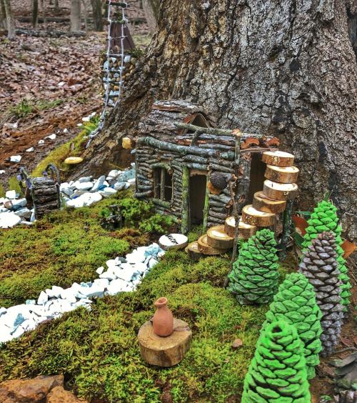 A whimsical miniature log cabin nestled at the base of a tree, surrounded by moss, stones, and tiny pine trees.