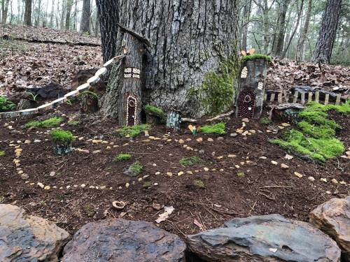 A whimsical miniature village made of natural materials, nestled at the base of a tree in a forested area.