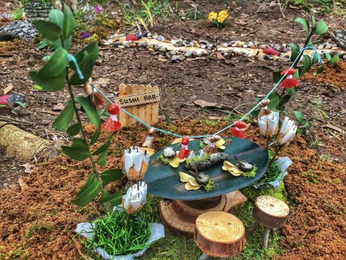 A whimsical miniature sushi bar scene with flowers, leaves, and decorative elements set in a natural outdoor setting.