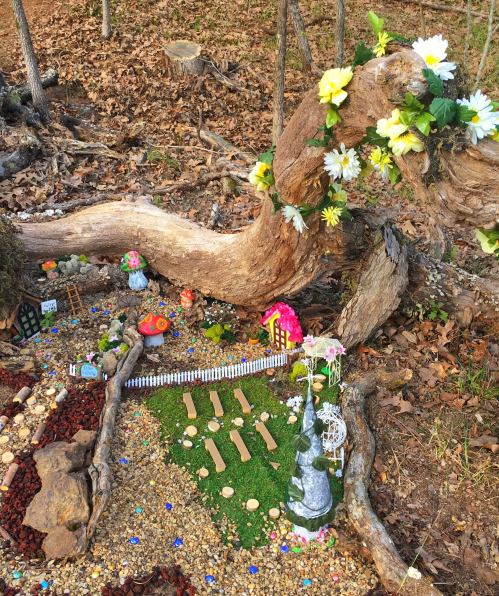A whimsical miniature garden scene with flowers, tiny houses, and decorative elements nestled among natural wood and stones.