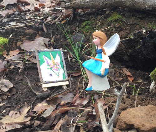 A small fairy figurine sits beside an easel with a colorful painting in a forest setting, surrounded by leaves and twigs.