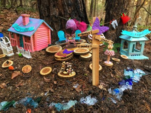 A whimsical miniature scene in the woods featuring colorful houses, signs, and decorative elements among tree roots.