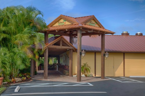 A welcoming entrance to a building with a brown roof, wooden beams, and tropical plants surrounding the pathway.