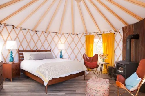 Cozy yurt interior featuring a bed, armchairs, a small table, and a wood stove, with warm lighting and decorative accents.