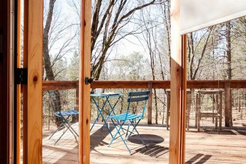 A view through wooden doors of a deck with blue chairs and a table, surrounded by trees in a serene outdoor setting.